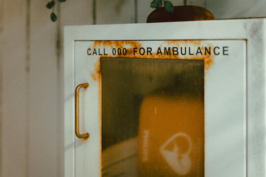 White metal cabinet mounted on a wall, labelled clearly with the text &ldquo;CALL 000 FOR AMBULANCE&rdquo;