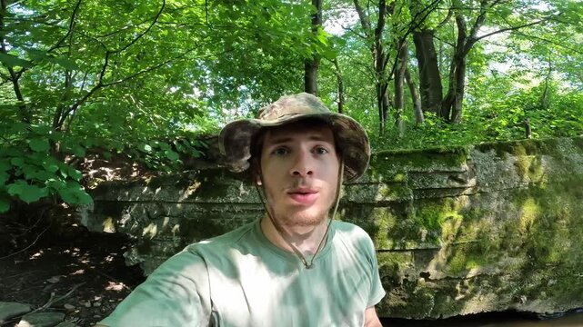 Outdoor content creator vlogging in front of an old mossy stone wall in a green summer forest, fishing lifestyle.
