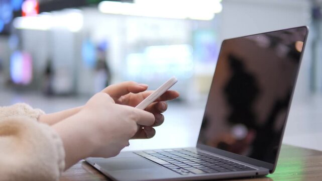 Person uses phone while laptop sits open nearby. Background shows public space with blurred lights and people. Hands hold phone close to screen. Soft lighting illuminates scene. Focus stays on hands