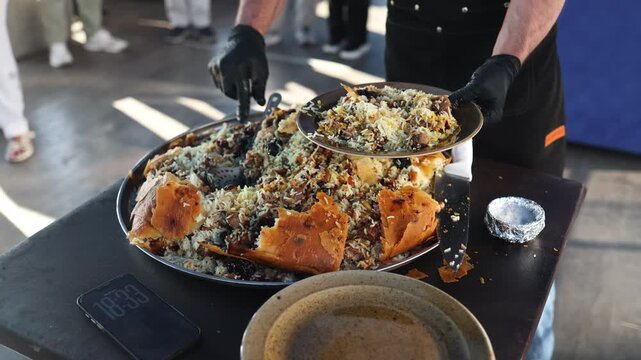 Cook pours delicious pilaf from large cauldron into bowl. Serving hot rice dish for traditional meal. Golden Uzbek pilaf served from cauldron into plate. Hearty rice dish prepared for nutritious meal.