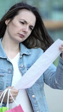 Young shopaholic woman with colorful bags shocked checking long receipt after shopping, worried about expenses and overspending.