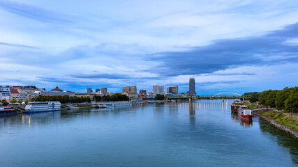Fototapeta premium Bratislava skyline across Danube River at dusk. Moored boats line both banks as city lights reflect beneath layered blue evening clouds.
