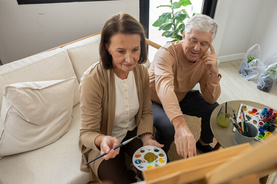 Senior couple painting art home canvas brush palette hobby creativity living room relaxed senior couple painting on canvas with brush and palette in cozy living room during creative hobby