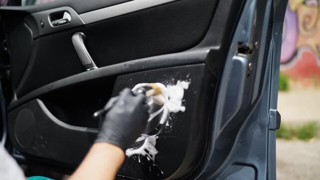 Professional auto detailer wearing black gloves scrubbing a dirty car door panel with a specialized detailing brush and applying cleaning foam for deep vehicle cleaning and restoration.