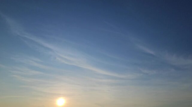 This stunning 4K video captures a tranquil sky during either sunrise or sunset, featuring a bright golden sun positioned near the horizon beneath a vast expanse of deep blue sky.