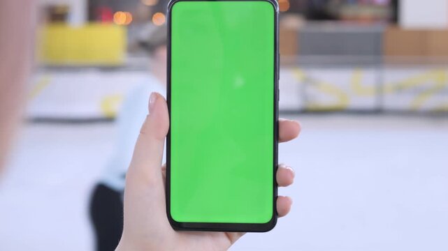 Hand holds phone with green screen, blurred indoor rink background. Person focused, calm, using device for content or video. Ideal for app demos, social media, or advertising visuals