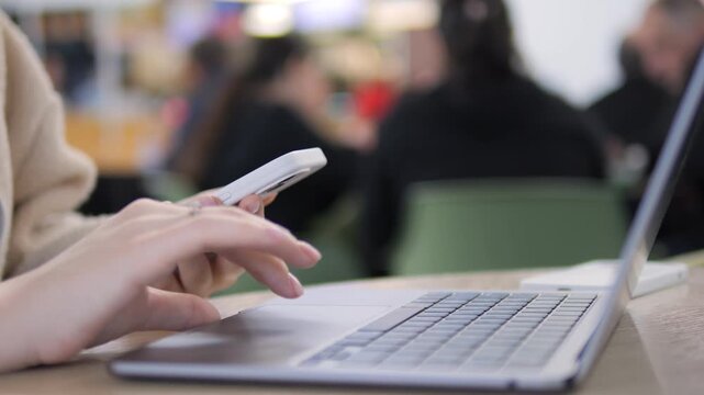 Close-up of hands using smartphone beside laptop. Person focused on phone while laptop remains open. Background shows blurred indoor space with people. Ideal for tech, multitasking