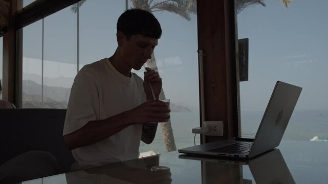 Location independent lifestyle with coastal remote work. Young freelancer taking coffee break while working on laptop at seaside cafe, digital nomad enjoying productive workday with ocean view in trop