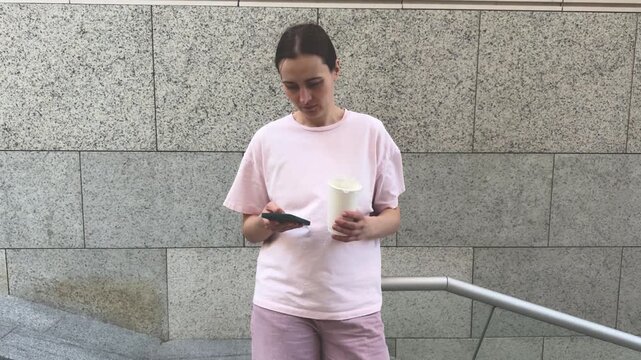 Young woman in pink outfit using smartphone while holding coffee cup against gray stone wall. Urban lifestyle, mobile technology, coffee break, casual fashion