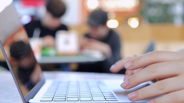 Hands type on laptop in cafe. Blurred background shows people working. Focus on keyboard and fingers. Suitable for remote work, study, or digital lifestyle themes
