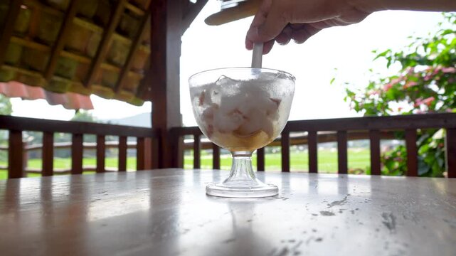 A glass of iced young coconut dessert with syrup being stirred on a wooden table.