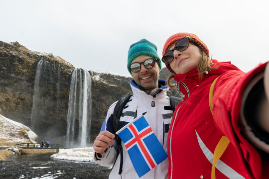 Iceland couple selfie wearing Icelandic sweaters by Seljalandsfoss waterfall on Ring Road in beautiful nature landscape on Iceland. Woman and man model in typical Icelandic sweater. Multiracial couple