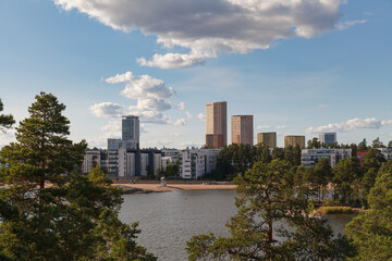 Obraz premium High-rise skyline of Helsinki in Finland viewed across a calm bay lined with pine trees and a sandy beach.