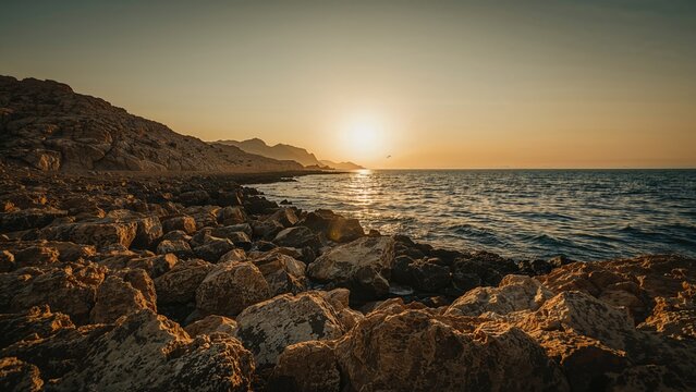 Sunset over a rocky coastline with the sun near the horizon and calm ocean waves.