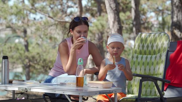 A mother and her young son enjoy a cozy picnic at a small camping table. The relaxed woman drinks warm tea while her boy eats bread. They share a peaceful moment in a sunny forest.