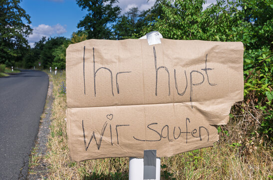 Handmade cardboard roadside sign with handwritten message mounted on wooden post beside quiet country road, near grassy verge, hedge, trees, sunlight, and open blue sky