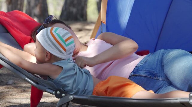 A happy mother and son relax together on a folding chair. They snuggle peacefully beside a blue camping tent in the quiet woods.