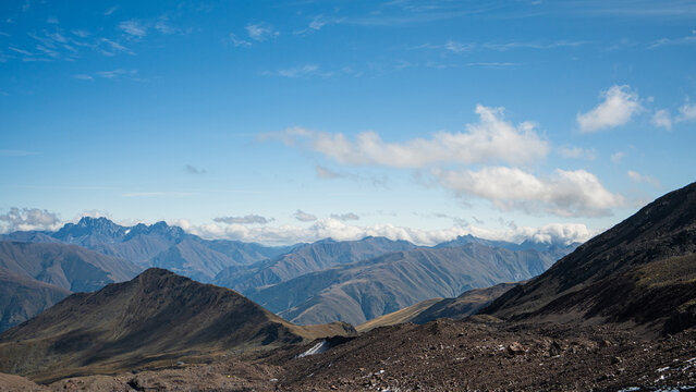 A panoramic view of layered mountain ridges receding toward distant jagged peaks under a blue sky with scattered clouds, Georgia.