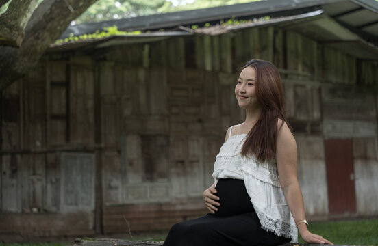 Pregnant woman in a white top smiles while sitting outdoors in front of a weathered rustic wooden building.