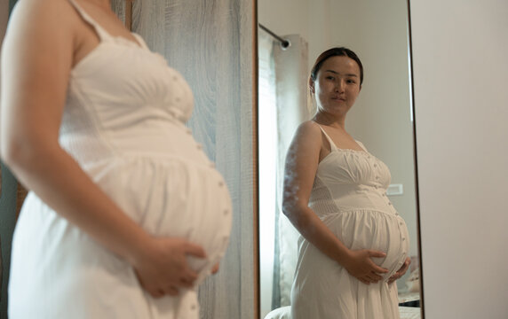 Pregnant woman in a white dress gently holds her belly while looking at her reflection in a bedroom mirror.