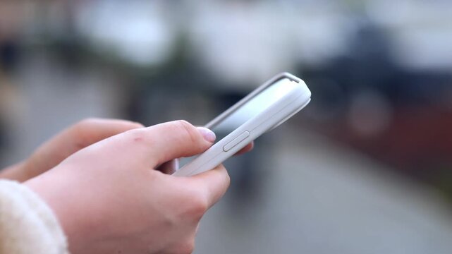 Close-up hands holding and scrolling smartphone outdoors. Focus on fingers interacting with screen. Background blurred with urban street setting. Person appears focused and engaged