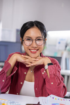 Woman eyeglass smile office desk shirt young Asian portrait workplace woman smiling with hands chin desk modern office, wearing eyeglass, pink shirt, and smartwatch, cheerful workplace Vertical