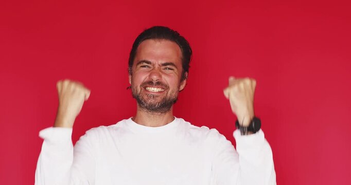 Handsome bearded man wearing white t shirt clenching fists shouting with powerful victorious expression and extreme excitement celebrating success with wide open mouth isolated red studio background