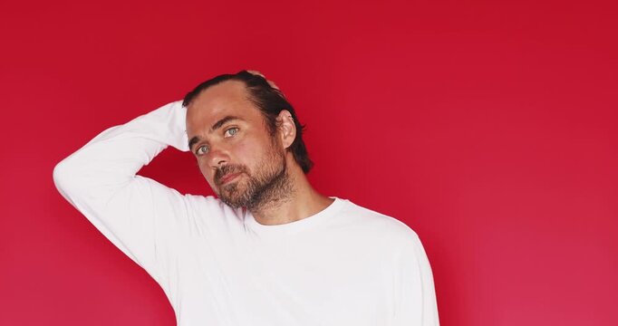 Handsome bearded man wearing white t shirt looking at camera and smiling with friendly facial expression after touching hair isolated on a solid red studio background close up portrait