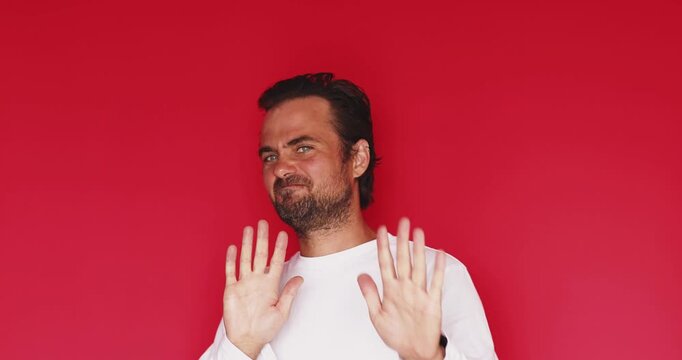 Handsome bearded man wearing white t shirt looking at camera and showing stop hand gesture with skeptical and rejecting facial expression while refusing something isolated on a solid red studio