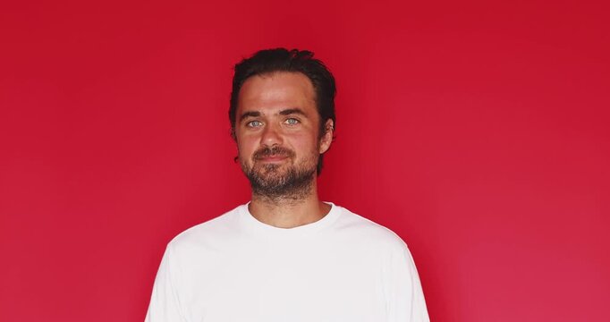 Handsome bearded man wearing white t shirt looking at camera and gesturing with skeptical facial expression while shaking head to say no and refusing something isolated on solid red studio background