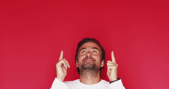 Excited middle aged Caucasian man looking up and pointing both index fingers up with amazed facial expression on vibrant red studio background, showing copy space area for text banner advertising