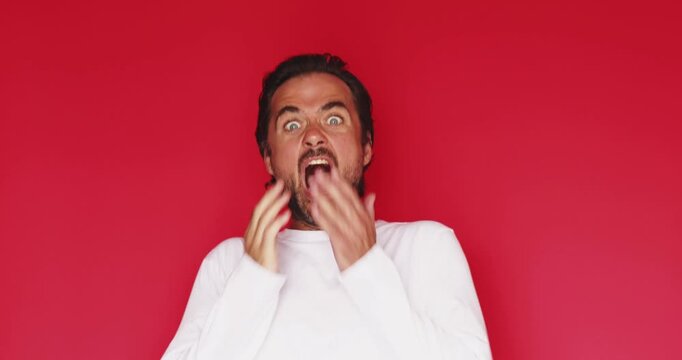 Middle aged Caucasian man wiping invisible glass or mirror with sleeve then covering mouth in shock and fear on vivid red studio background, funny mistake and cleaning surprise conceptual stock video