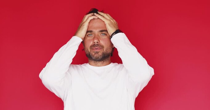Shocked middle aged Caucasian man holding head in hands with open mouth from stress then breathing out with calm serious expression on bright red studio background, panic to relief concept