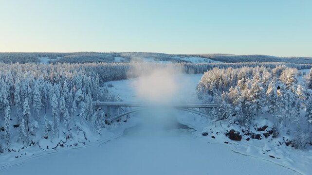 Aerial drone view of a bridge over a river in a snowy pine forest on a foggy winter day. Misty seasonal forest scene.