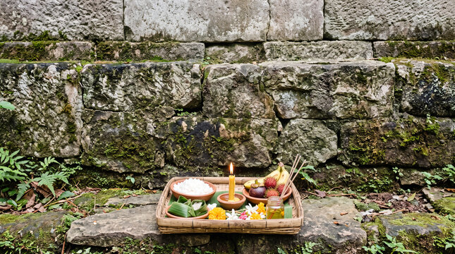 A woven offering tray of fruit, rice, flowers and a lit candle placed at the foot of a mossy stone wall