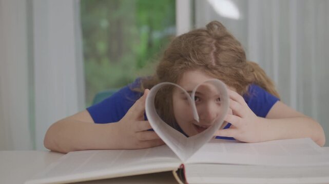 Happy Child Making Heart Shape with Book Pages. School child behind an open book with pages folded into a heart shape. Learning, reading bools, education, imagination, love of books concept.