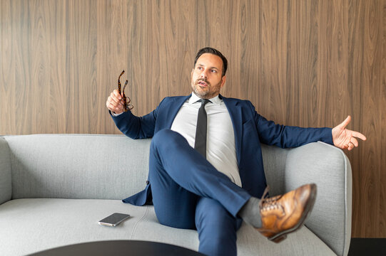 Confident man relaxing on a studio couch, holding glasses, and discussing ideas while looking away, reflecting leadership and business success