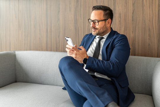 Businessman sitting on office seating, focused on his mobile device, engaging in digital communication and managing tasks while maintaining a professional demeanor
