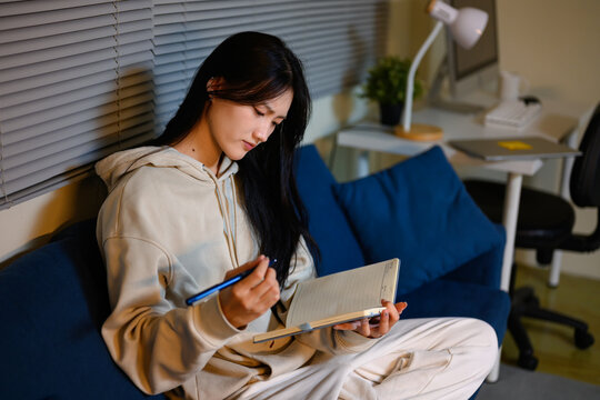 Young Asian Woman Writing in Notebook on Sofa at Home at Night in Cozy Room