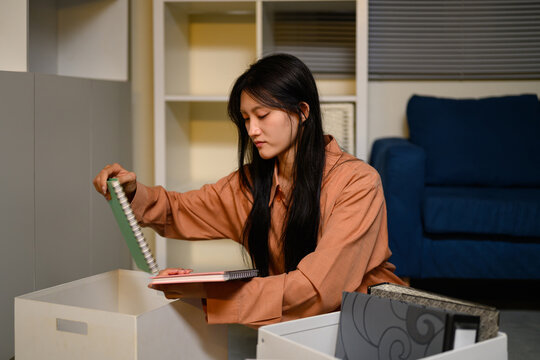 Asian businesswoman packing personal belongings into box after resignation, clearing office desk with notebooks and files