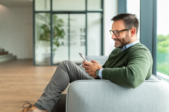 Smiling man relaxing on a gray upholstered seating, typing on his mobile device, enjoying connectivity and digital communication in a contemporary business setting