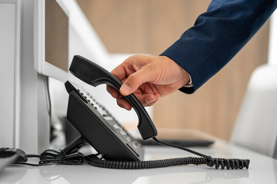 Businessman's hand picking up a telephone receiver from a desk phone, symbolizing communication, business contact, calling, and support in an office setting
