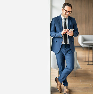 Business executive smiling while checking messages on his mobile phone, leaning against a wall during a break in a modern office environment, connecting with clients or colleagues