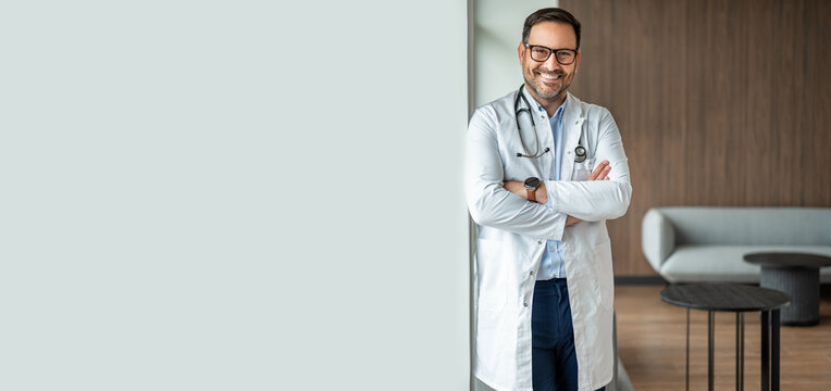 Confident male doctor in a medical uniform smiling with crossed arms standing in a clinic reception, presenting medical care, health support, and consultation services