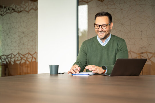 Smiling professional man working at a modern office desk, actively engaging with his laptop and notebook while planning and managing business projects