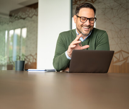 Professional businessman wearing glasses smiling and gesturing during a video conference, actively engaging in virtual meeting discussions using a laptop at his office workspace