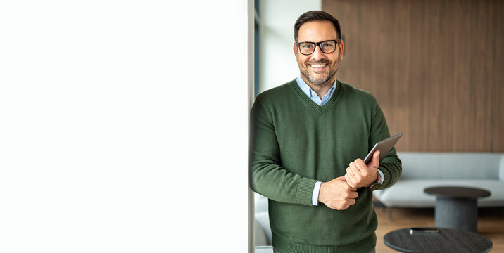 Confident male executive smiling, holding a tablet, posing against a modern office background with room for text, symbolizing innovation and corporate success