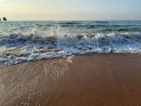 A spilling wave bursts into bright sea foam on a sandy shore under cool silver-white light. Textured water and shimmering reflections capture the moment where the ocean meets the beach.