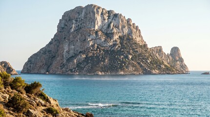 Naklejka premium Es Vedra limestone rock island rises from the Mediterranean sea near Ibiza. The scene captures a peaceful morning atmosphere with clear blue water. Ideal for travel brochures and nature blogs.