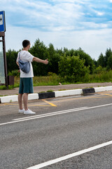 Man with a bag hitchhikes on a rural road by extending his thumb. The wide road stretches into the distance with a vehicle approaching, highlighting travel and adventure themes. © magpie